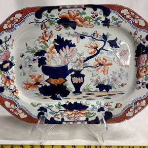 May include: An oval ceramic platter featuring a white base with a detailed floral pattern in dark blue, orange, and green. The border has a decorative orange and blue design. A yellow measuring tape shows the platter is 15 inches in length.