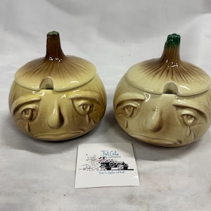 Peut inclure: Deux pots en céramique en forme d'oignon avec couvercle et finition brillante, arborant un visage triste avec des marques en forme de larmes, un couvercle à tige brune et une encoche pour cuillère. Une carte blanche avec Fat Cats Antiques.