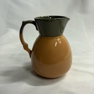 May include: A ceramic pitcher featuring a rounded, light tan body and a dark olive green upper section and handle. The two colors are separated by a subtle, raised beaded texture that encircles the neck of the vessel, which has a spout.