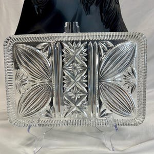 May include: A rectangular clear glass serving tray featuring a pressed pattern with a central vertical column of starburst motifs flanked by large fan-shaped designs. The edges are finished with a repeating vertical ribbed texture.