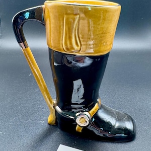 May include: A ceramic mug shaped like a tall black riding boot with a tan cuff at the top. The handle is black and extends from the top rim to the heel. A decorative tan strap with a silver-toned buckle detail wraps around the ankle area.