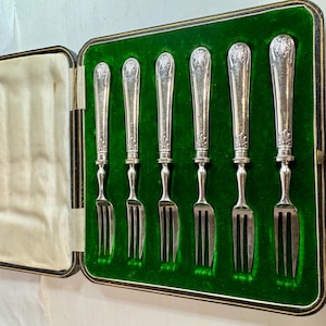 Könnte beinhalten: Ein Set aus sechs silberfarbenen Metallgabeln mit verzierten, geprägten Griffen, die an der Oberseite ein muschelartiges Motiv aufweisen. Die Gabeln sind vertikal in einem rechteckigen, aufklappbaren Etui mit grünem Samtfutter angeordnet.