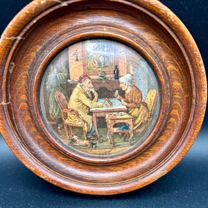 May include: A circular wooden frame containing a colored print of two people sitting at a table playing cards. The scene includes a small dog on the floor, a person standing in the background, and the text A PAIR printed at the bottom center.