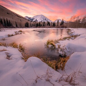 Puede incluir: Un paisaje nevado con un lago congelado que refleja los tonos rosados y anaranjados de una puesta de sol. Una cordillera nevada se eleva en el fondo, con una línea de árboles a lo largo del borde del lago.