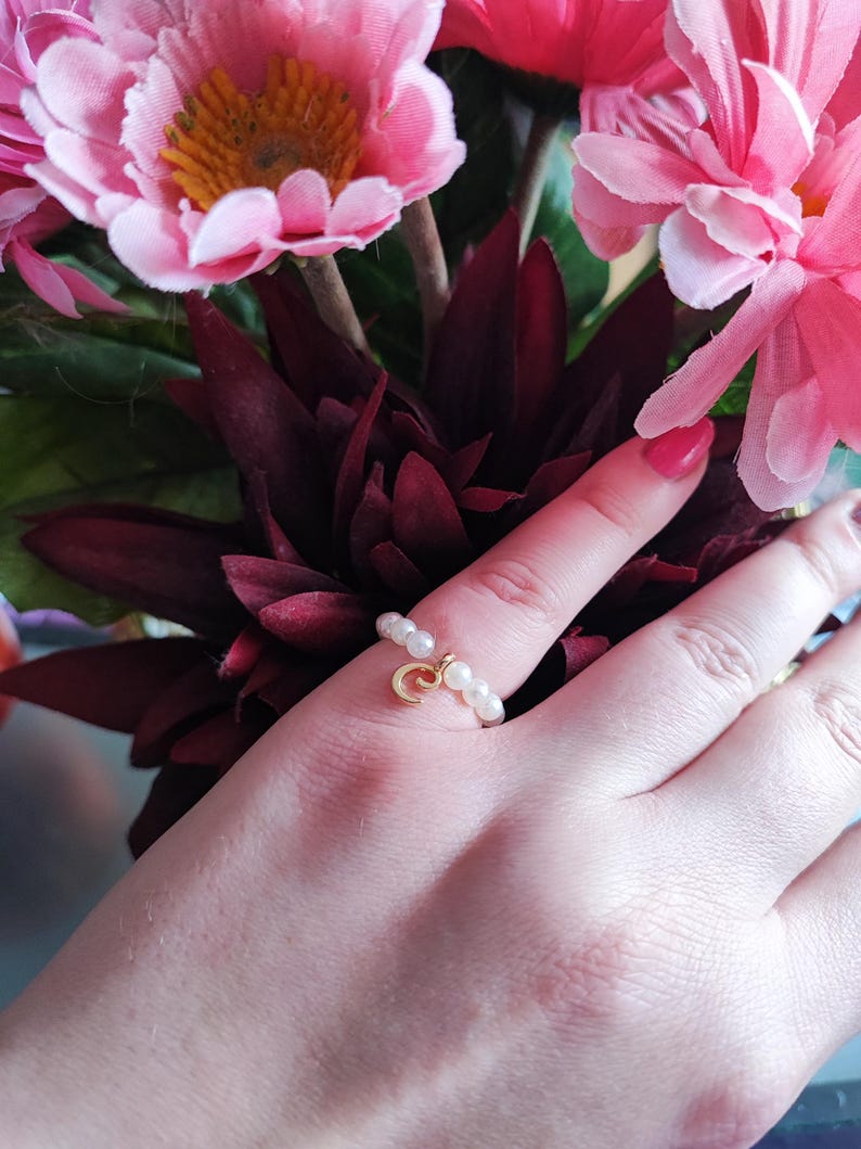 Może przedstawiać: Złoty pierścionek z zawieszką w kształcie p&oacute;łksiężyca i małymi białymi perłami. Pierścionek jest na palcu na tle ciemnoczerwonej i r&oacute;żowej kompozycji kwiatowej.