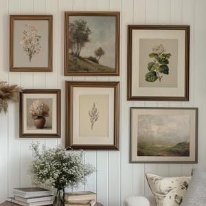 May include: A gallery wall arrangement featuring six framed prints with dark brown frames, including botanical illustrations and landscape paintings, displayed above a wooden side table with books, a glass vase of flowers, and a white sofa.