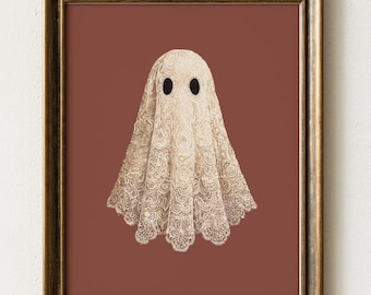 May include: A framed wall art print featuring a central illustration of a ghost figure draped in intricate, cream-colored lace fabric. The ghost has two solid black oval eyes and is set against a solid, muted terracotta-colored background.
