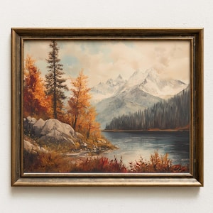 May include: A landscape painting in a dark gold frame depicting a calm lake surrounded by autumn trees with orange and yellow foliage, rocky shores, and snow-capped mountains under a cloudy sky. The artwork captures a serene mountain scene.