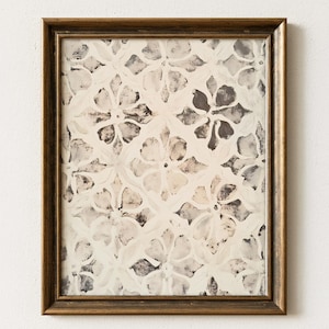 May include: A framed artwork featuring a floral pattern in shades of white, cream, and gray. The design is repeated across the surface, creating a textured effect. The frame is a warm, golden brown color, adding a touch of elegance to the piece.