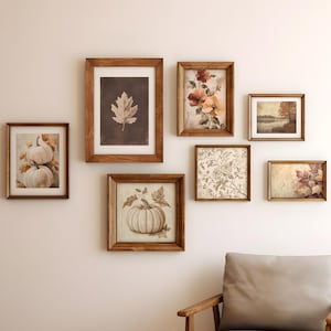 May include: A gallery wall arrangement featuring seven framed prints in wooden frames. The artwork includes depictions of white pumpkins, a single oak leaf on a dark background, orange flowers, a landscape scene, floral patterns, and autumn foliage.