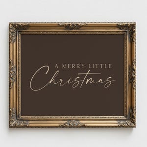 May include: A rectangular wall art print in a dark brown frame with ornate gold detailing. The print features a dark brown background with the text A MERRY LITTLE Christmas written in a combination of serif and script fonts in a light beige color.