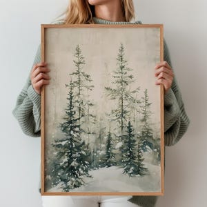 May include: A rectangular framed wall art print featuring a watercolour painting of a snowy forest scene. The artwork depicts several evergreen pine trees in shades of dark green and muted grey against a soft, pale beige background with white snow.
