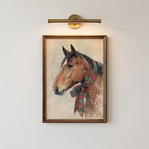 May include: A framed wall art print featuring a brown horse wearing a red and green plaid scarf, depicted against a light background with white specks resembling falling snow, illuminated by a brass-toned horizontal wall light fixture above.