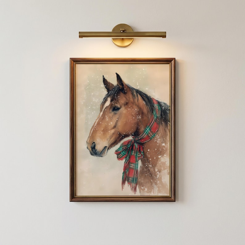 May include: A framed wall art print featuring a brown horse wearing a red and green plaid scarf, depicted against a light background with white specks resembling falling snow, illuminated by a brass-toned horizontal wall light fixture above.