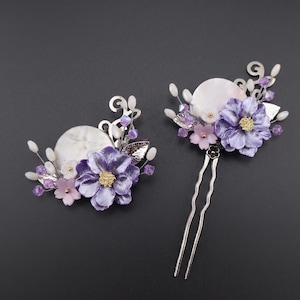 May include: Two floral hair accessories. One is a barrette, the other a hair pin. Both feature a central white disc, surrounded by purple and pink fabric flowers, silver leaves, and small purple beads. The background is a dark gray.