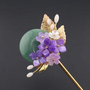 May include: A decorative hair stick featuring a green jade disc, adorned with purple and white flower accents, gold leaves, and a gold-colored stem. The floral arrangement is detailed and elegant.