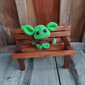 May include: A handmade crochet figure of a character with large green ears, a brown robe, and black eyes. The figure is sitting on a miniature wooden bench with a brown finish. The background is a wooden wall.