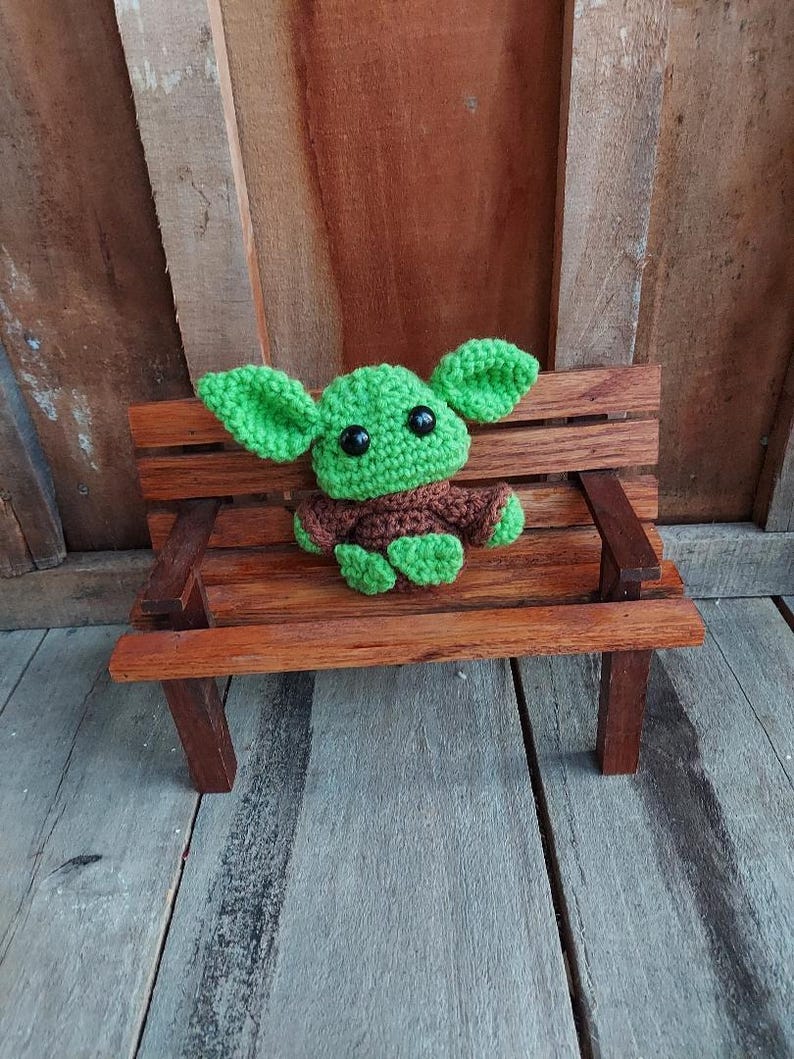 May include: A handmade crochet figure of a character with large green ears, a brown robe, and black eyes. The figure is sitting on a miniature wooden bench with a brown finish. The background is a wooden wall.