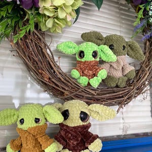 May include: A collection of crocheted plush toys, each resembling a well-known character. They have large black eyes, green skin, and are dressed in brown or tan outfits. The toys are set against a floral wreath.