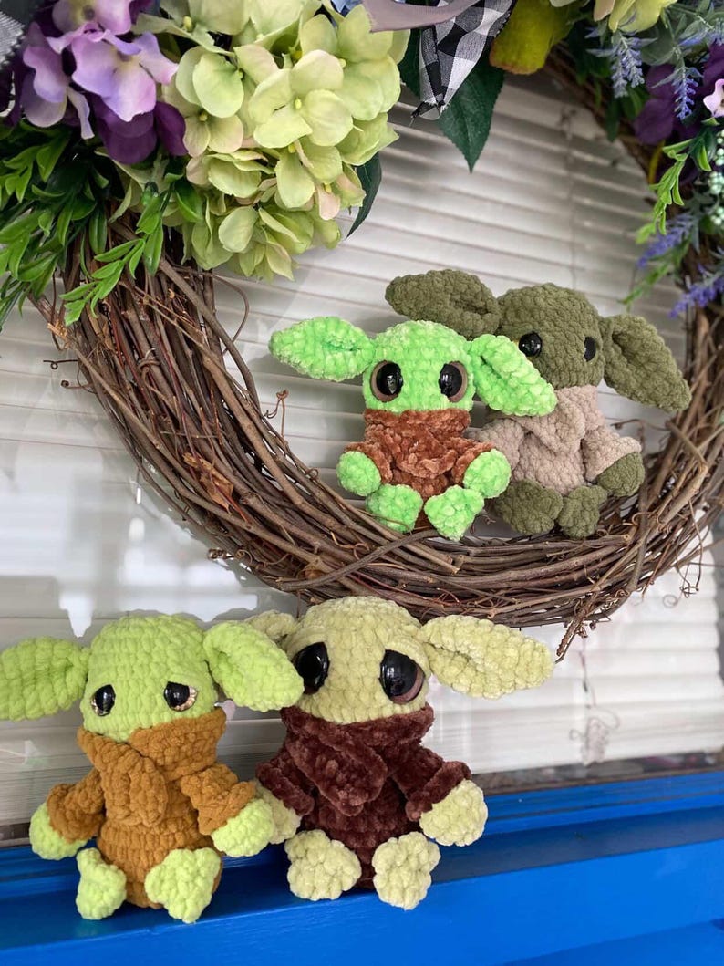May include: A collection of crocheted plush toys, each resembling a well-known character. They have large black eyes, green skin, and are dressed in brown or tan outfits. The toys are set against a floral wreath.