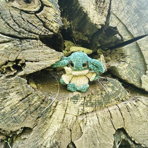 May include: A handcrafted, green and beige crocheted toy with large, dark eyes and pointed ears. The plush figure is placed within the textured surface of a weathered tree stump. The toy's design is inspired by a well-known character from a popular science fiction series.