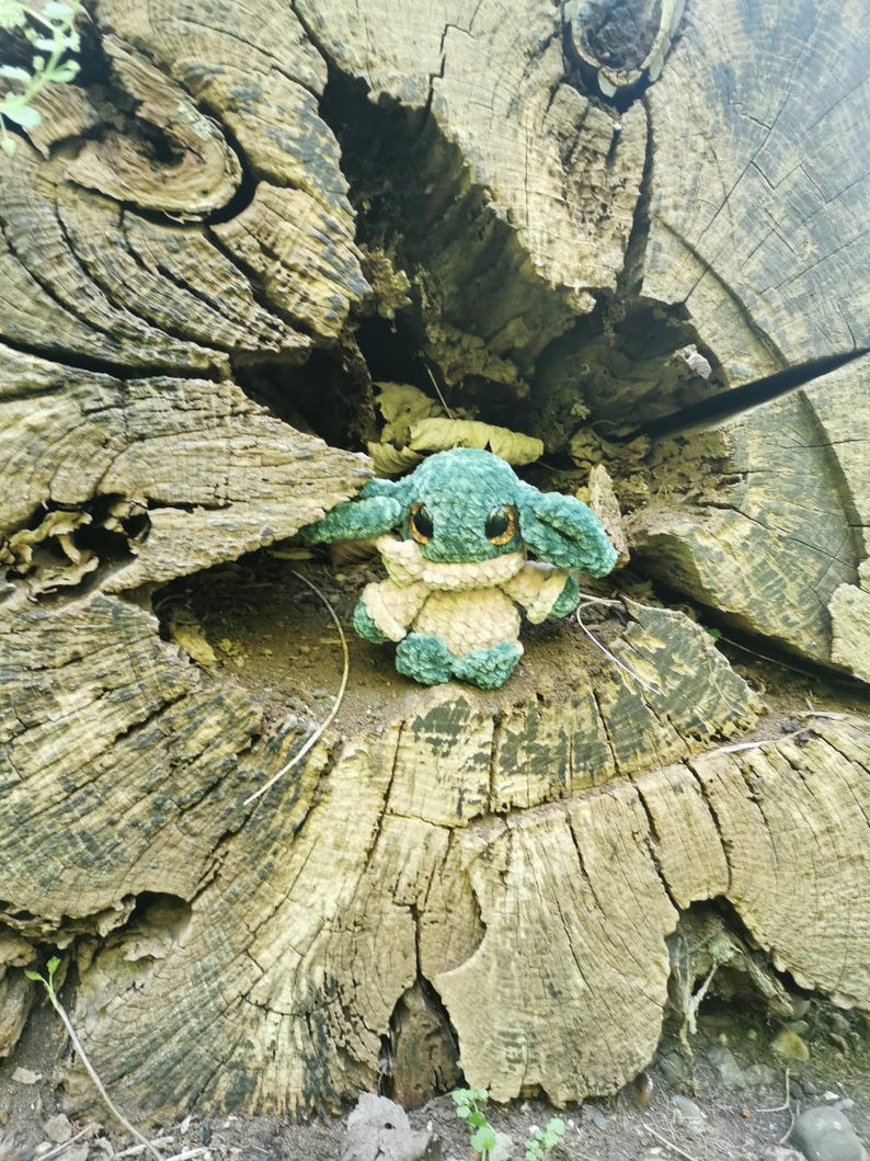 May include: A handcrafted, green and beige crocheted toy with large, dark eyes and pointed ears. The plush figure is placed within the textured surface of a weathered tree stump. The toy's design is inspired by a well-known character from a popular science fiction series.