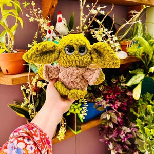 May include: A crocheted doll resembling a well-known character, with large black eyes, oversized ears, and a tan and green colour scheme. The doll is held in front of a shelf with plants and decorative items.