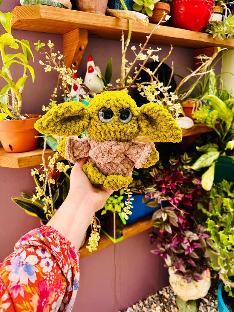 May include: A crocheted doll resembling a well-known character, with large black eyes, oversized ears, and a tan and green colour scheme. The doll is held in front of a shelf with plants and decorative items.