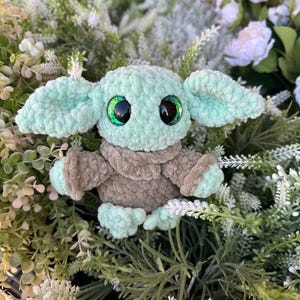 May include: A handmade crochet doll resembling a popular character. The doll has large, expressive eyes with green highlights, large ears, and a soft, plush texture. The colour scheme includes shades of mint green and brown.