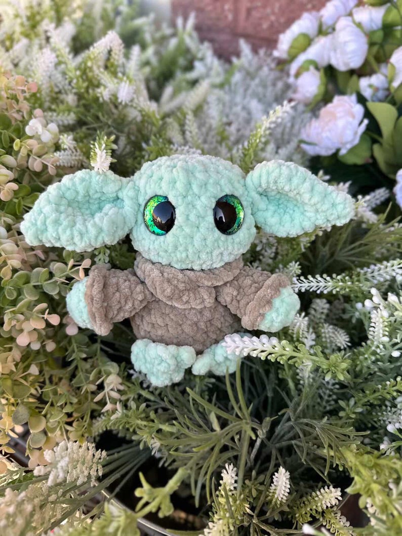 May include: A handmade crochet doll resembling a popular character. The doll has large, expressive eyes with green highlights, large ears, and a soft, plush texture. The colour scheme includes shades of mint green and brown.