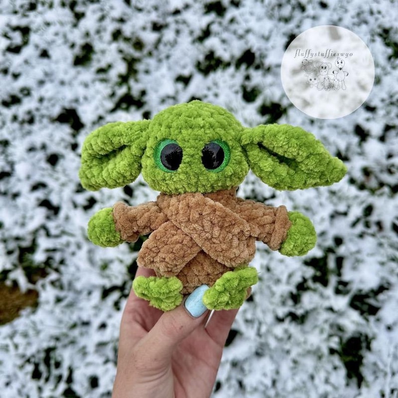 May include: A handmade green and brown plush doll with large black eyes and oversized ears. The doll is wearing a brown robe and has green hands and feet. The doll is sitting in front of a snowy background.