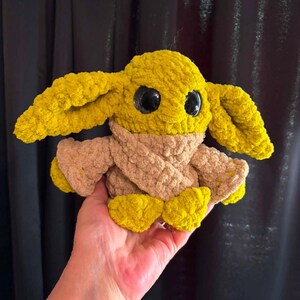 May include: A crocheted plush toy resembling a character from a science fiction franchise. The toy has large black eyes, yellow ears, and a tan scarf. The body and feet are yellow. The toy is held in a hand.