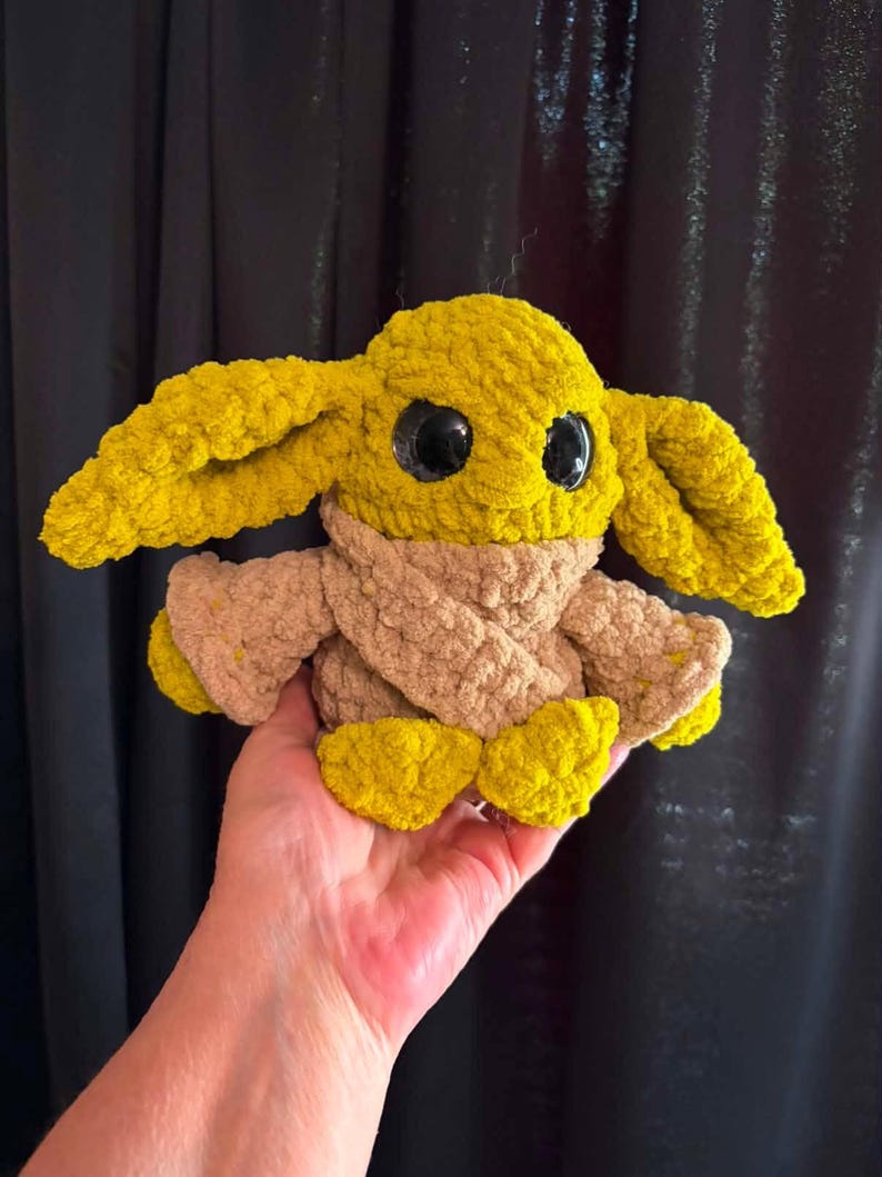 May include: A crocheted plush toy resembling a character from a science fiction franchise. The toy has large black eyes, yellow ears, and a tan scarf. The body and feet are yellow. The toy is held in a hand.