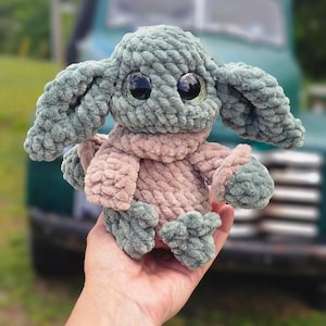 May include: A handmade plush toy resembling a well-known character, with large, expressive eyes. The toy is mainly green and tan, with a soft, textured appearance. The toy is held in a hand, with a blurred background of a vintage truck.