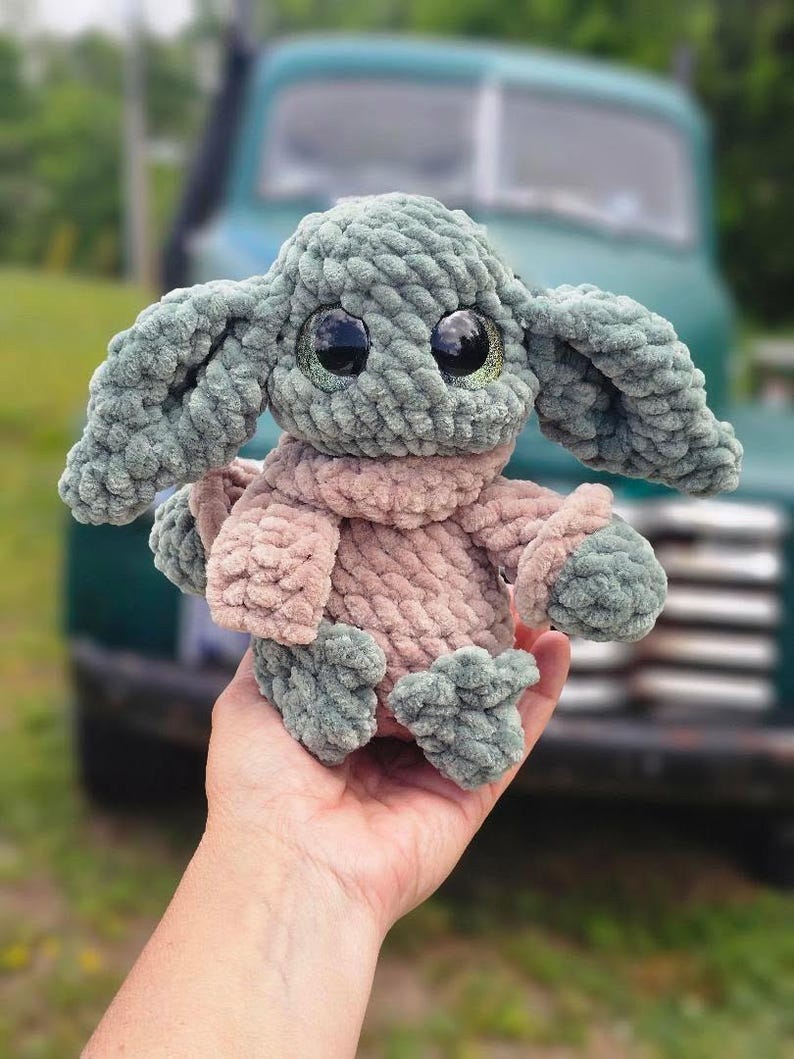 May include: A handmade plush toy resembling a well-known character, with large, expressive eyes. The toy is mainly green and tan, with a soft, textured appearance. The toy is held in a hand, with a blurred background of a vintage truck.
