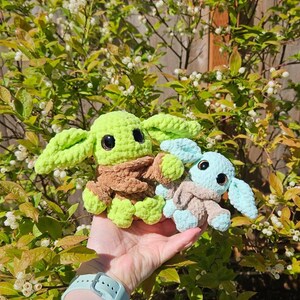May include: Two crocheted plush figures, one green and brown, the other blue and tan, resembling a popular character. The figures have large black eyes and oversized ears. The background features green foliage and white flowers.