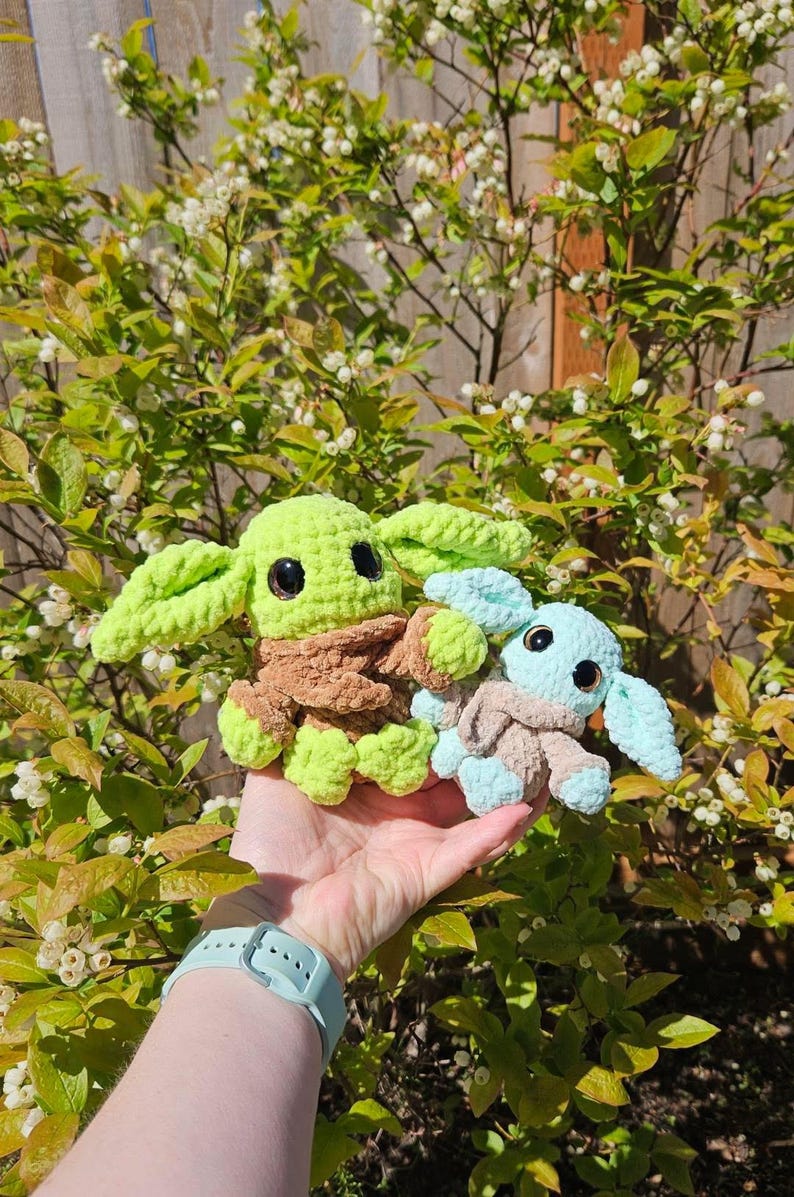 May include: Two crocheted plush figures, one green and brown, the other blue and tan, resembling a popular character. The figures have large black eyes and oversized ears. The background features green foliage and white flowers.