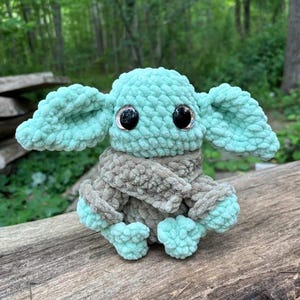 May include: A crocheted plush toy resembling a character with large ears and big black eyes. The toy is mainly mint green and grey, with a textured, fuzzy appearance. It is sitting on a weathered wooden surface.