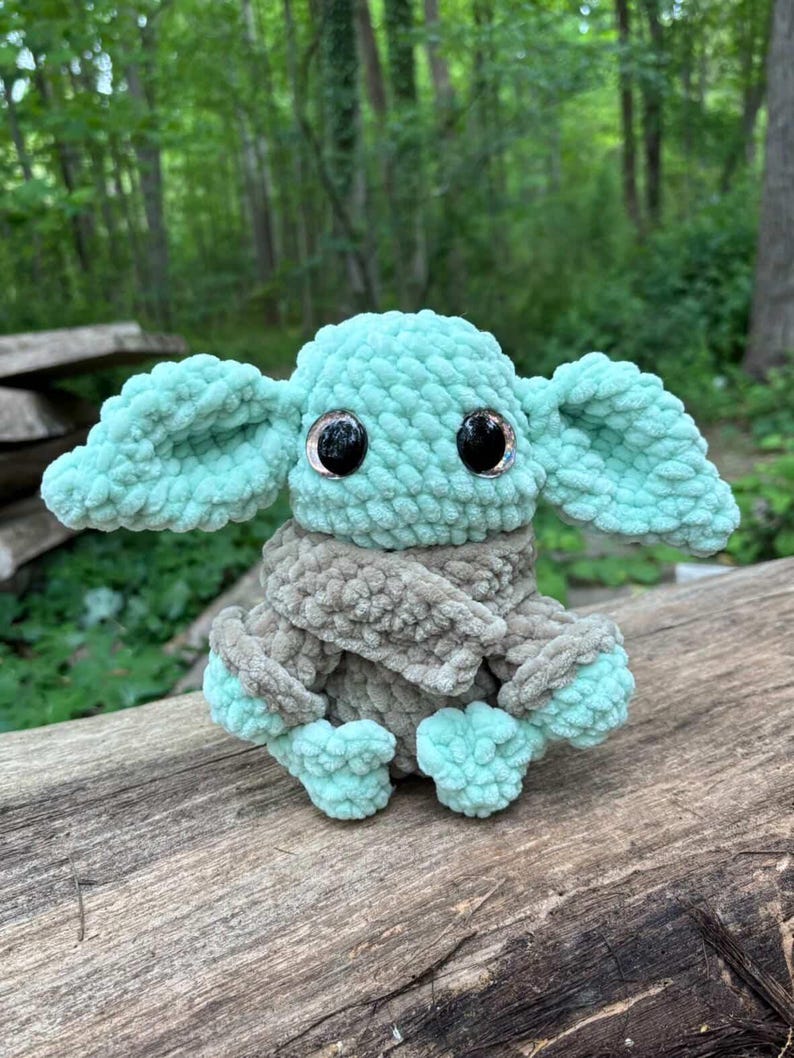 May include: A crocheted plush toy resembling a character with large ears and big black eyes. The toy is mainly mint green and grey, with a textured, fuzzy appearance. It is sitting on a weathered wooden surface.