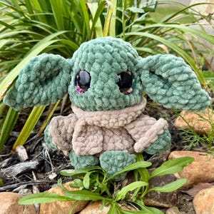 May include: A handmade plush doll resembling a well-known character. The doll is mainly green with large, black eyes and a beige scarf. The doll is made of a soft, textured material and is set amongst green foliage.