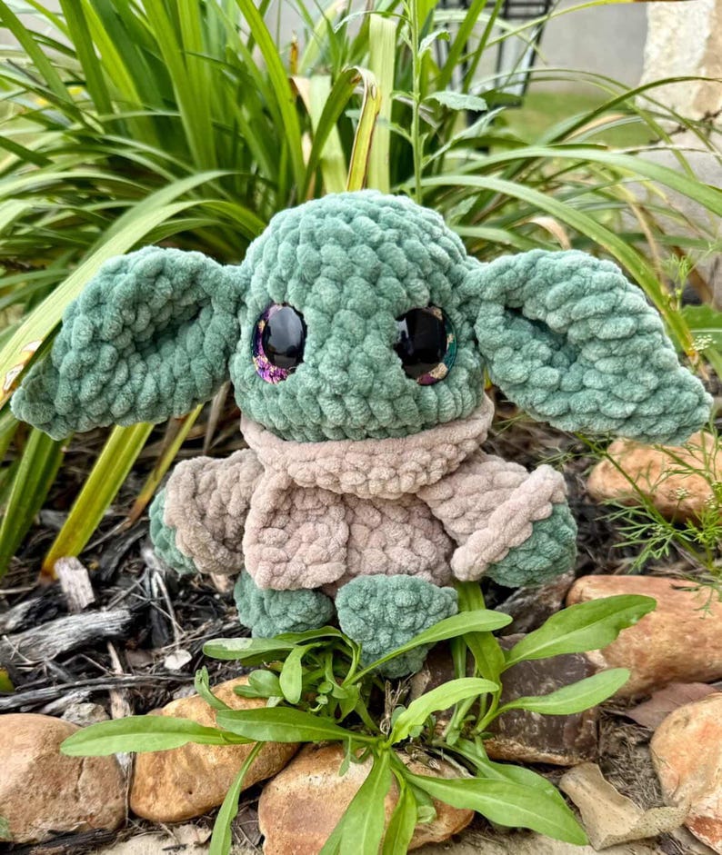 May include: A handmade plush doll resembling a well-known character. The doll is mainly green with large, black eyes and a beige scarf. The doll is made of a soft, textured material and is set amongst green foliage.