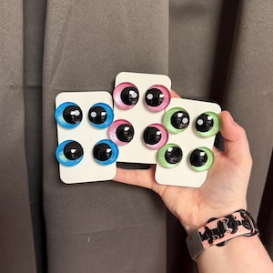 May include: A collection of six pairs of glass eye cabochons in various colors, including blue, pink, and green, each with a black pupil. The eyes are mounted on white cardstock. The image is held by a person wearing a watch.