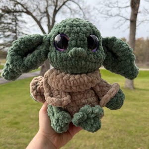 May include: A handmade green and brown crocheted plush toy with large, black eyes and purple irises. The toy is wearing a brown coat with green trim and has large, floppy ears. The plush is held in a person's hand.