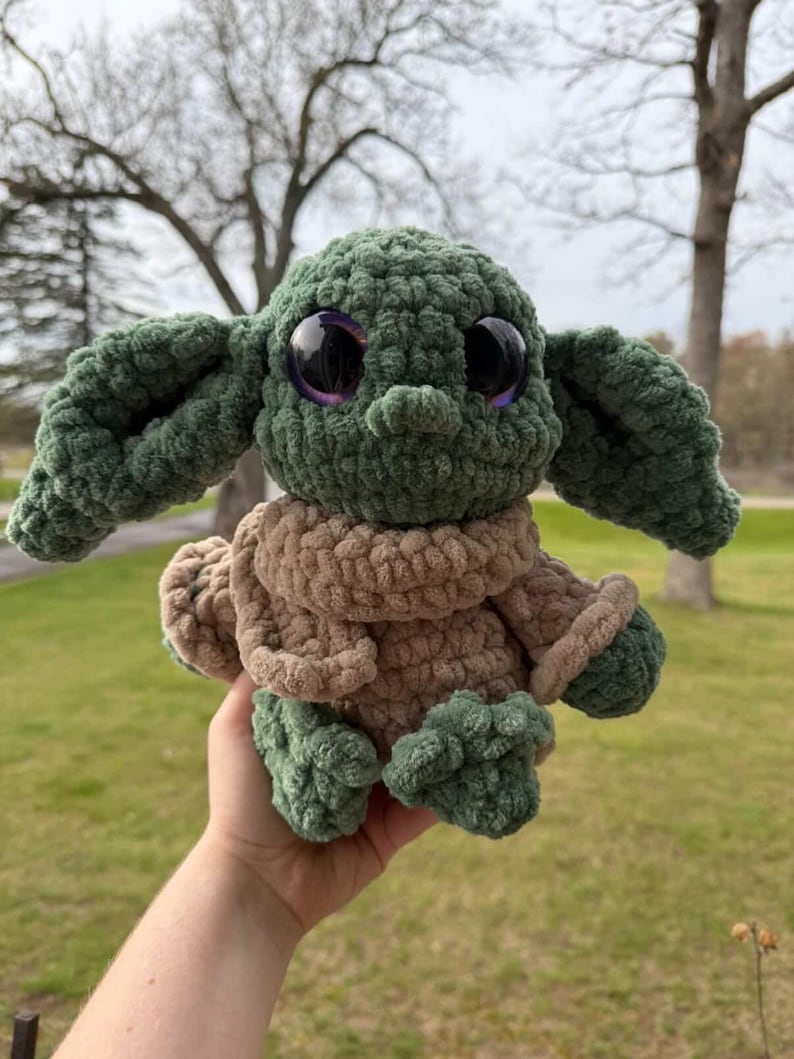 May include: A handmade green and brown crocheted plush toy with large, black eyes and purple irises. The toy is wearing a brown coat with green trim and has large, floppy ears. The plush is held in a person's hand.
