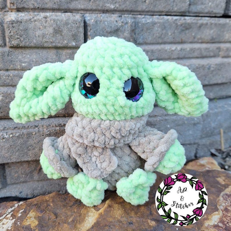 May include: A crocheted plush toy resembling a character with large eyes and ears. The toy is mainly light green with a grey scarf and feet. The eyes are black with iridescent details. A circular logo with text is in the lower right corner.