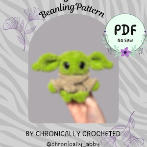 May include: A digital PDF pattern titled Baby Alien Beanling Pattern by Chronically Crocheted. The image features a small, green, plush alien figure with large ears and dark eyes, wearing a tan wrap, with text indicating it is a no-sew project.