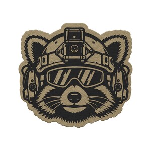 May include: A tan and black patch featuring a raccoon wearing tactical gear, including a helmet, goggles, and headset. The raccoon's face is detailed with black fur and a small nose. The patch has a textured, leather-like appearance.
