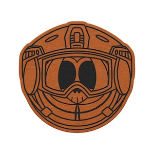 May include: A round, tan leather patch featuring a black line drawing of a cartoon-style helmet with goggles. The helmet has large, black eye shapes and a smiling mouth. The patch is likely for apparel or accessories.
