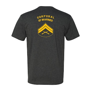 May include: A dark heather grey t-shirt with the text "Corporal of Marines" and a yellow chevron with crossed rifles.