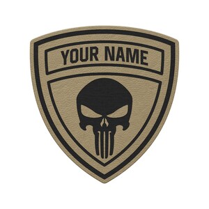 May include: A tan shield-shaped patch with a black border and a black skull graphic in the center. The upper section of the patch features the text YOUR NAME in bold black capital letters, set against a textured tan background.
